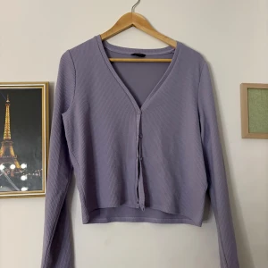 Lila ribbad kofta - Säljer en stilren lila kofta med ribbad struktur och v-ringning. Koftan har långa ärmar och knappar framtill, perfekt för lager-på-lager-stil.