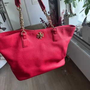 Röd axelväska från Tory Burch - Snygg röd axelväska med guldfärgad kedja och logotypdetalj framtill. Perfekt för att ge en färgklick till din outfit. Väskan har en rymlig design och är lätt att bära med sig.