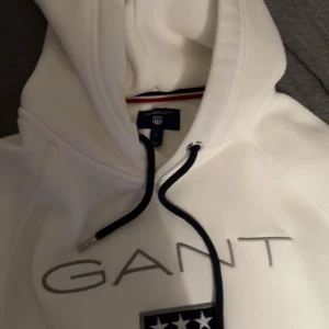 Vit hoodie från GANT - Säljer en stilren vit hoodie från GANT med logotyp och ett broderat emblem på bröstet. Hoodien har en justerbar dragsko i huvan och en praktisk magficka. Perfekt för en avslappnad stil.