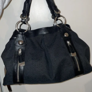 Svart handväska från DKNY - Snygg svart handväska från DKNY med diskret mönster och silverfärgade detaljer. Väskan har två dragkedjor på framsidan och stora ringar som fäster handtagen. Insidan är rymlig med flera fack och en dragkedja för extra säkerhet. Har lite fläckar på tyget på insidan (se sista bilderna)