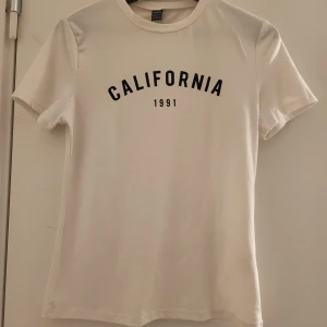 Vit t-shirt med tryck 'California 1991' - Snygg vit t-shirt med texten 'California 1991' tryckt i svart på framsidan. Perfekt för en avslappnad stil. Kortärmad och i mjukt material. Använt 1 gång. Köpt för 100 kr från shein säljer för 80kr