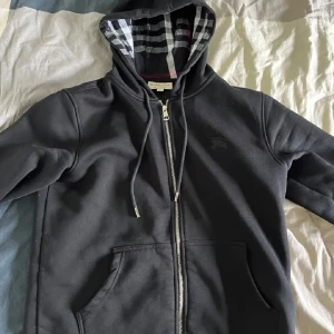 Svart huvtröja med dragkedja - Snygg svart huvtröja från Burberry med dragkedja och klassiskt rutigt mönster i huvan. Perfekt för en stilren och bekväm look. Tröjan har långa ärmar och praktiska fickor framtill. Hade tröjan uppe ett tag men valde att inte sälja den, men nu är det dax. Den är i mycket bra skick