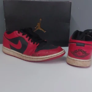 Nike Air Jordan 1 Low i röd och svart - Säljer ett par Nike Air Jordan 1 Low i röd och svart. Skorna har en klassisk design med snörning och en ikonisk logga på hälen. Perfekta för sneakerheads som älskar retrostil.