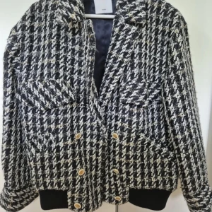 Svartvit rutig jacka från Sandro - Snygg svartvit rutig jacka från Sandro med dubbelknäppning och stora fickor framtill. Jackan har en klassisk krage och är perfekt för att ge en stilren look. Den är långärmad och har en bekväm passform.