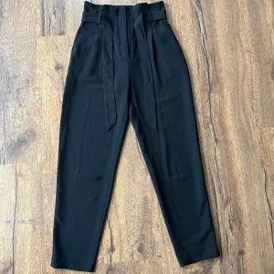 Svarta kostymbyxor med knytband - Snygga svarta kostymbyxor med hög midja och knytband i midjan. Byxorna har en dragkedja och knapp framtill även fickor som är diskreta. Även ”fejk” fickorna bak på byxorna. Använda men inget tecken på slitage. Säljs för de inte kommer till användning😍