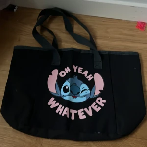 Svart shoppingväska med tryck stitch  - Snygg svart shoppingväska med ett roligt tryck av en tecknad figur och texten 'Oh Yeah Whatever'. Väskan har två rejäla handtag och är perfekt för att bära med sig allt man behöver. Tillverkad i ett slitstarkt material.