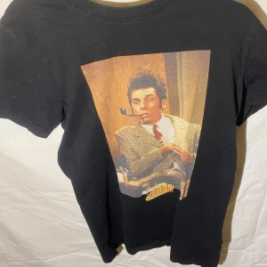 Svart Seinfeld t-shirt från Dedicated - Cool svart t-shirt från Dedicated med ett tryck av en karaktär från Seinfeld. Tillverkad i ekologisk bomull och Fairtrade-certifierad. Perfekt för fans av serien! 