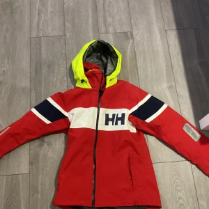 Röd jacka från Helly Hansen salt 2 - Säljer en röd jacka från Helly Hansen med en iögonfallande gul huva och marinblå detaljer. Jackan har en dragkedja framtill och långa ärmar med vita och blå ränder. Perfekt för kyliga dagar. OBS katt finns i hemmet pris kan diskuteras