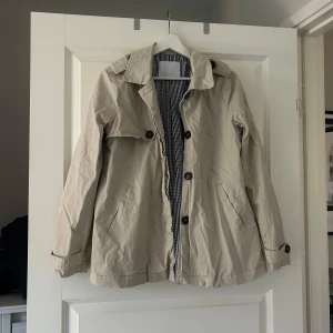 Beige trenchcoat med knappar - Snygg beige trenchcoat med knappar och krage. Jackan har långa ärmar och två fickor framtill. Perfekt för vår och höst. Beige kappa kort kappa kort trenchcoat 