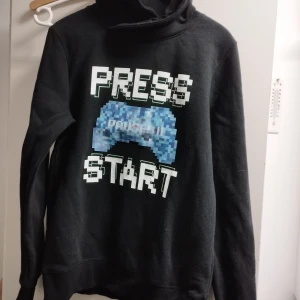 Svart hoodie med spelmotiv från H&M - Cool svart hoodie från H&M med texten 'PRESS START' och en pixlad spelkontroll i blått och vitt på framsidan. Perfekt för gaming-entusiaster! Tröjan har långa ärmar och en bekväm passform. Barn/ungdom 