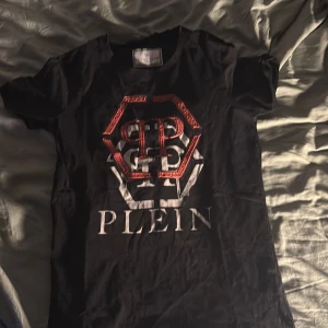 Svart t-shirt från Philipp Plein Junior - Snygg svart t-shirt från Philipp Plein Junior med ett stort rött och vitt tryck på framsidan. Perfekt för en cool och avslappnad stil. T-shirten har korta ärmar och en klassisk rund halsring, tyvär finns det 2 pytte små hål som knappt syns ens när du har på den. 