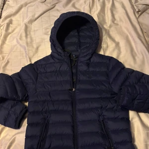 Polo ralphlauren dunnjacka  - Snygg mörkblå dunjacka från Polo Ralph Lauren med huva och dragkedja framtill. Jackan har två praktiska fickor med dragkedjor och är perfekt för kyligare väder.