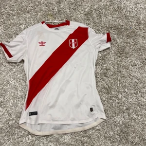 Peru fotbollströja - Säljer en snygg vit fotbollströja från Umbro med ett diagonalt rött band och ett emblem på bröstet. Tröjan har korta ärmar och en klassisk rund hals. Perfekt för fotbollsfans!