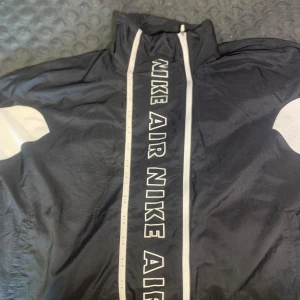Svart vindjacka från Nike Air (ST L) - Snygg svart vindjacka från Nike Air med vita detaljer och logga på både fram- och baksidan. Jackan har en dragkedja framtill och är perfekt för en sportig look. Tillverkad i lätt och bekvämt material.