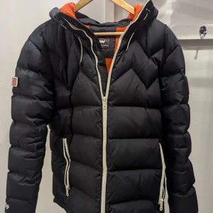 Svart dunjacka Mountain Works - Säljer en snygg svart Dunjacka från Mountain Works med coola orange detaljer. Jackan har en dragkedja framtill och är perfekt för kalla dagar. Den har en skön huva.  Perfekt för vintern och hösten.