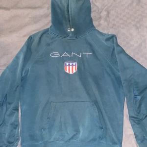 Grön hoodie från GANT - Säljer en snygg grön hoodie från GANT med deras ikoniska logga och sköld på bröstet. Perfekt för kyliga dagar med sin mjuka och bekväma passform. Den har en klassisk känguruficka. Passar både till vardags och avslappnade tillfällen.