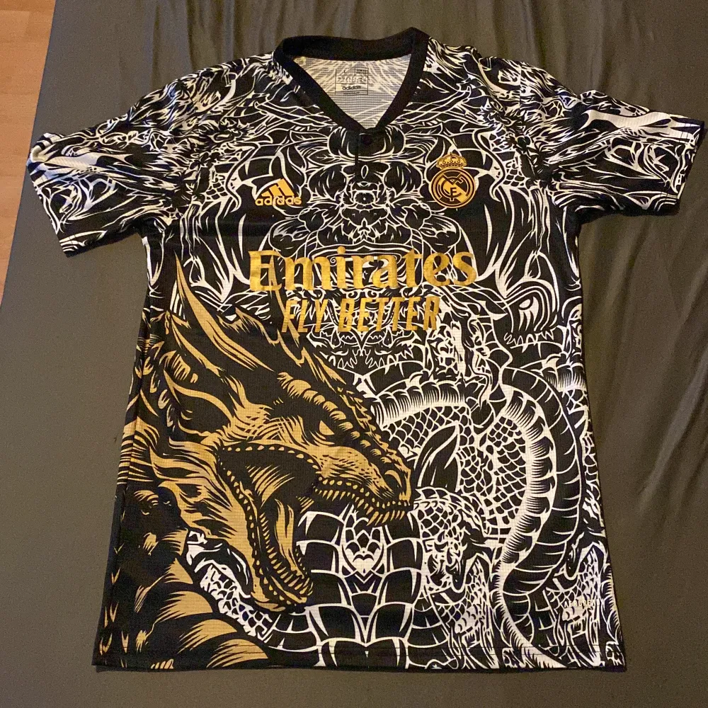 Säljer en  svart och guld Real Madrid tröja från Adidas. Den har ett episkt drakmönster i vitt och guld som täcker hela tröjan. Perfekt för att sticka ut på matchen eller bara ha som en snygg streetwear-stil. Kortärmad och i mycket bra skick! Priset går att diskutera . T-shirts.