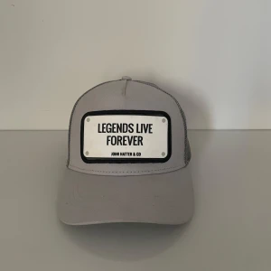 Grå keps från John Hatter & Co - Snygg grå keps med texten 'Legends Live Forever' på framsidan. Märket John Hatter & Co är broderat under texten. Kepsen har en klassisk design med justerbar baksida i mesh för bra ventilation. Perfekt för både vardag och fest! Den är köpt i butik och tillkommer utan box.              Tveka inte att kontakta om du har frågor eller funderingar😁