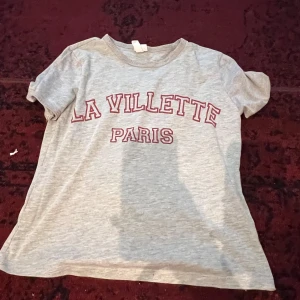 Grå t-shirt med text 'La Villette Paris' - Säljer en grå t-shirt med röd text 'La Villette Paris' på framsidan. Den är kortärmad och i normal passform, perfekt för en avslappnad stil. Materialet känns mjukt och bekvämt, idealiskt för vardagsbruk.