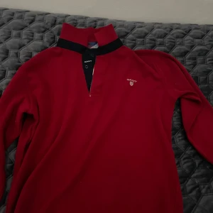 Röd tröja från GANT - Säljer en snygg röd tröja från GANT i bra skick. Den har en stilren design med en liten logga på bröstet och en krage med knappar. Perfekt för både vardag och lite finare tillfällen. Passar bra till höst och vinter.