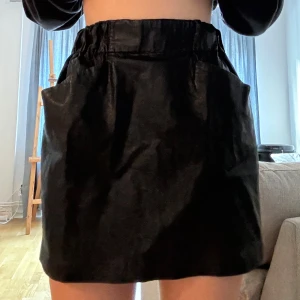 Svart kjol i skinnimitation - Snygg svart kortkjol i skinnimitation med hög midja och elastisk resår. Perfekt för en kväll ute eller en trendig vardagslook. Den har en enkel design utan extra detaljer, vilket gör den lätt att matcha med olika toppar och accessoarer.