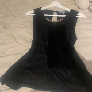 Svart ärmlös klänning med metallisk detalj - Snygg svart ärmlös topp med en cool metallisk detalj vid halsen. Perfekt för en kväll ute eller en fest. Toppen har en figursydd passform och är gjord i ett mjukt material som faller fint.