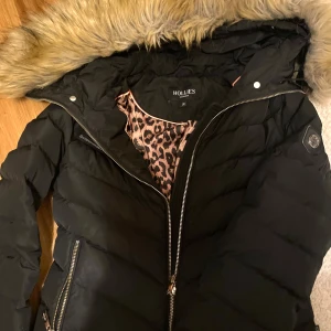 Svart dunjacka från Hollies - Säljer en snygg svart dunjacka från Hollies i storlek S/36 . Jackan har en mysig pälskantad huva och ett coolt leopardmönstrat foder. Perfekt för kalla vinterdagar med dragkedja och knappar för extra värme. 🖤