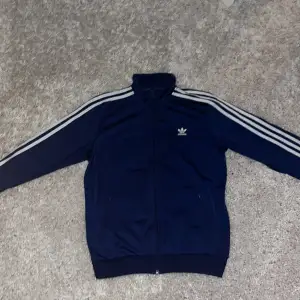 Säljer en snygg mörkblå tröja från Adidas med de klassiska tre ränderna längs ärmarna. Jackan har en dragkedja framtill och ett stort Adidas-logotyp på ryggen. Den är i mycket bra skick och redo för nya äventyr!