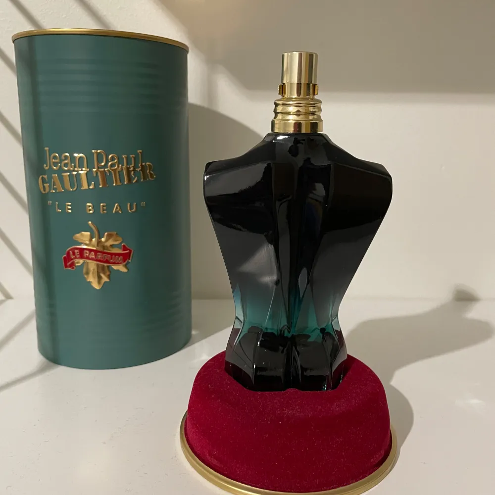 Säljer en cool parfymflaska formad som en manlig torso från Jean Paul Gaultier. Flaskan är mörkgrön och svart med en guldig spraytopp. Den kommer i en elegant grön metallförpackning med guldtext. Perfekt för den som vill ha en unik och stilren doftupplevelse. Flaskan står på en röd sammetsbas.. Perfume.
