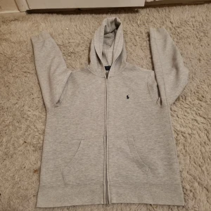 Grå hoodie från Polo Ralph Lauren - Säljer en snygg och bekväm grå hoodie från Polo Ralph Lauren. Den har en dragkedja framtill och en klassisk logga på bröstet. Perfekt för både vardag och chill. Den är i bra skick och passar till alla tillfällen!