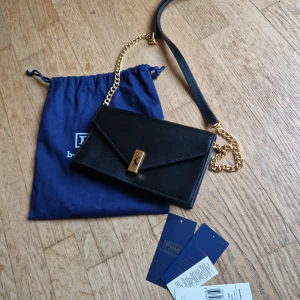 Svart axelväska från Polo Ralph Lauren - Snygg svart axelväska i skinn från Polo Ralph Lauren. Väskan har en elegant guldkedja och ett stilrent spänne framtill. Perfekt för både vardag och fest! Den kommer med en blå dustbag för förvaring. Helt ny med tags kvar.