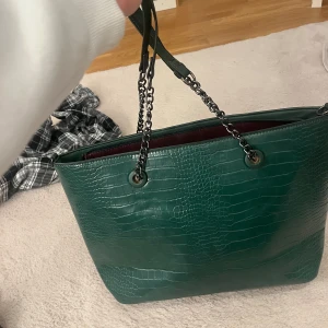 Grön axelväska med krokodilmönster - Snygg grön axelväska i syntetmaterial med krokodilmönster. Väskan har kedjehandtag och en dragkedja upptill. Perfekt för att ge en touch av stil till din outfit. Passar bra för både vardag och fest!