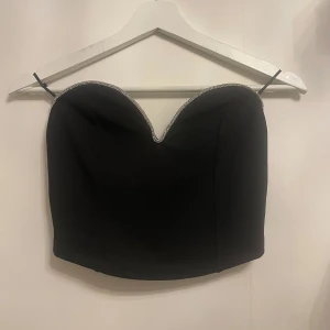 Svart axelbandslös topp med strassdetaljer - Snygg svart axelbandslös topp med glittrande strassdetaljer längs kanten. Perfekt för festliga tillfällen! Toppen har en tight passform och är croppad för en trendig look. Passar bra till både kjol och byxor. Perfekt till nyår 