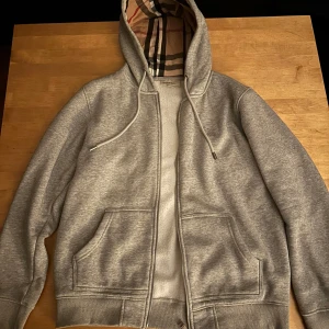 Grå burberry hoodie med dragkedja - Säljer en grå hoodie i bomull med dragkedja och en klassisk rutig insida i huvan. Perfekt för höst och vår, med långa ärmar och fickor framtill. Superbekväm och stilren för vardagsbruk! Storlek L men kan passa M