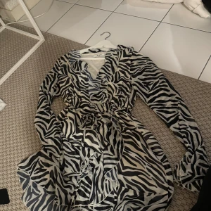 Zebramönstrad klänning från NA-KD - Zebramönstrad klänning från NA-KD i storlek 36. Klänningen har långa ärmar och en volangdetalj vid fållen samt knytning i midjan. Den är använd fåtal gånger, så i fint skick 