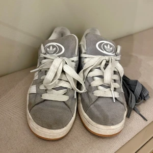 Grå Adidas sneakers Campus - Säljer ett par grå Adidas sneakers i bra skick. De har en klassisk design med vita snören och den ikoniska Adidas-loggan på plösen. Skorna är perfekta för vardagsbruk och har en bekväm passform. Extra skosnören medföljer.
