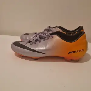 Säljer ett par grymma Nike Mercurial fotbollsskor i silver och orange. De har en snygg svart Nike-logga och snörning för perfekt passform. Perfekta för fotbollsspelare som vill ha stil och prestanda på planen. Skorna är i bra skick och redo för nya äventyr!