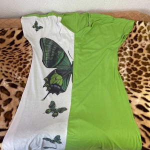 Grön och vit klänning med fjärilsmotiv - Säljer en unik klänning i grönt och vitt med ett stort fjärilsmotiv. Klänningen har en normal passform och är perfekt för sommardagar. Materialet känns mjukt och bekvämt mot huden. Perfekt för dig som vill sticka ut med ett naturinspirerat mönster!