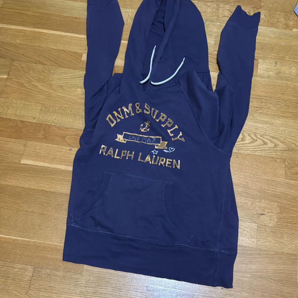 Säljer en snygg mörkblå hoodie från Ralph Lauren, perfekt för en avslappnad stil. Den har en stor ficka fram och en justerbar huva med snören. Texten 'DNM & SUPPLY' är tryckt i gult på framsidan. Passar perfekt för höst och vinter! Lila blå färg. Tröjor & Koftor.