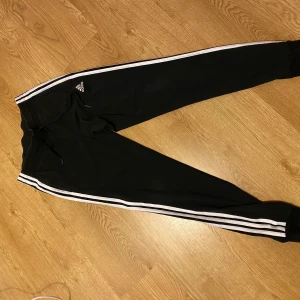 Svarta träningsbyxor från Adidas - Säljer ett par svarta träningsbyxor från Adidas i storlek S. Byxorna har vita ränder längs sidorna och en bekväm passform med resår i midjan. Perfekta för träning eller en avslappnad dag hemma. 🖤pris kan diskuteras.