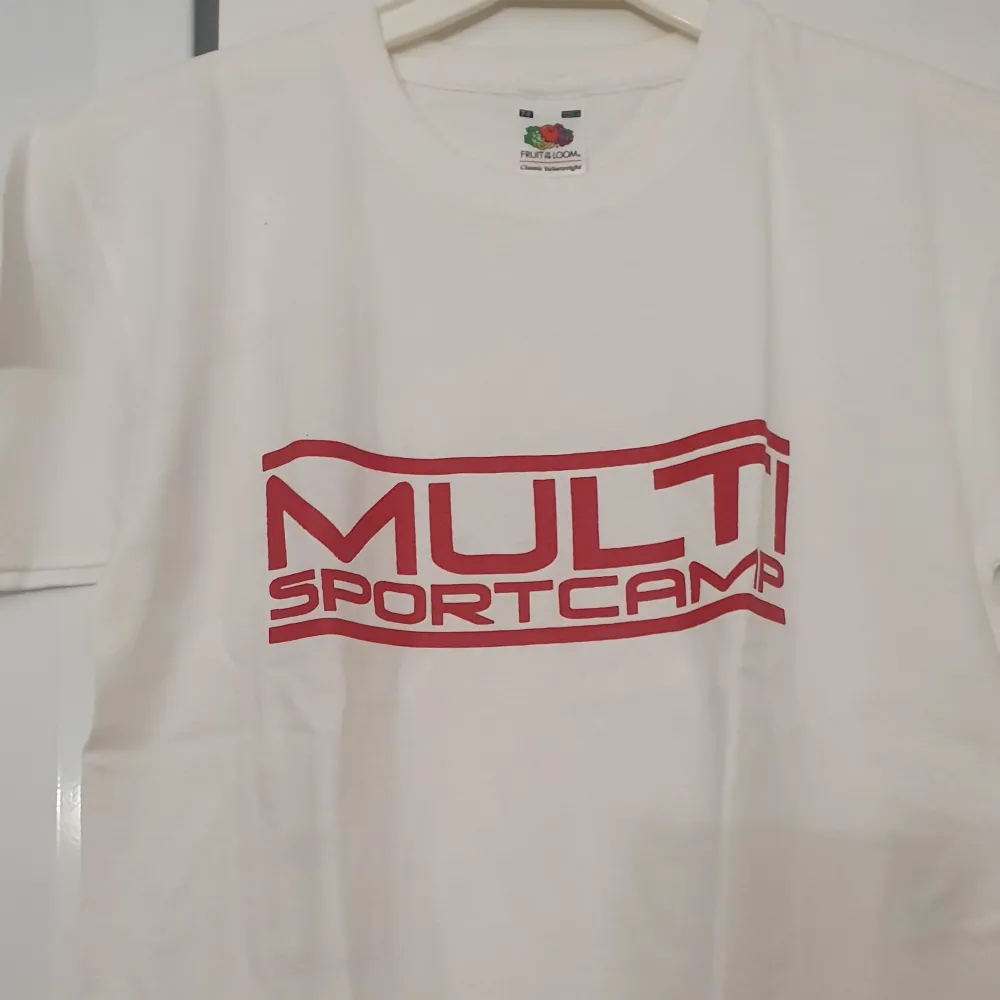 Säljer en vit t-shirt från Fruit of the Loom i klassisk stil. Den har en röd text 'MULTI SPORTCAMP' tryckt på framsidan. Perfekt för sportiga aktiviteter eller som en casual vardagströja. Materialet är mjukt och bekvämt, idealiskt för barn i åldern 7-8 år.. T-shirts.