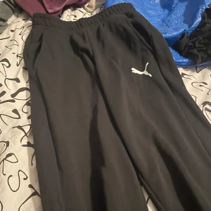Svarta mjukisbyxor från Puma - Säljer ett par svarta mjukisbyxor från Puma i bra skick. De har en elastisk midja och är perfekta för en avslappnad stil. Passar både för hemmamys och lätt träning. Puma-loggan syns tydligt på benet.
