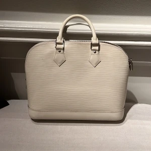 Benvit handväska från Louis Vuitton - Säljer en elegant benvit klassisk ”Alma” från Louis Vuitton i Epi-skinn. Väskan har en stilren design med dragkedja och ett LV-lås 