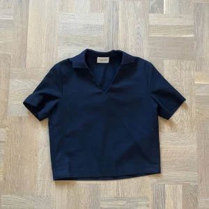 Mörkblå topp från Wood Wood - Säljer en stilren mörkblå topp från Wood Wood i storlek 36. Den har en enkel design med korta ärmar och en v-ringad krage. Perfekt för både vardag och lite mer uppklädda tillfällen.