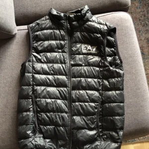 Svart dunväst från EA7 Emporio Armani - Säljer en snygg svart dunväst från EA7 Emporio Armani. Den är i bra skick enligt mig sen har en färg fläck på insidan av den man ser inte så mkt. Sen har den ett styng där bak men det ser man knappt. 