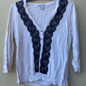 Vit tröja med spetsdetaljer från H&M - Säljer en superfin vit tröja från H&M med svarta spetsdetaljer längs med knäppningen. Tröjan har trekvartsärmar och en v-ringad design som ger en elegant touch. Perfekt för både vardag och fest! 🖤