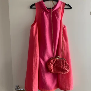 Klänning och väska - A-linje skuren klänning i fusia och korall. Köpt på HM i deras special sortiment. 87cm från Axel till nederkant. Stl.40 men upplevs som small. Linne 43% och Lycocell 57%. Mycket fint skick. Tillhörande aftonväska.