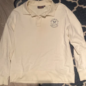 Vit pikétröja från Morris Stockholm - Säljer en stilren vit pikétröja från Morris Stockholm. Tröjan har en klassisk krage och ett tryck på bröstet med texten 'Morris Raquet & Tennis Sporting Club'. Perfekt för en avslappnad men ändå sportig look. Passar bra till både vardag och lite finare tillfällen.
