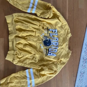 Gul tröja med Florida-tryck - Säljer en cool gul tröja med tryck av 'Florida' och en basketboll på framsidan. Tröjan har långa ärmar med vita ränder och slitna detaljer som ger en vintage look. Perfekt för en avslappnad stil!