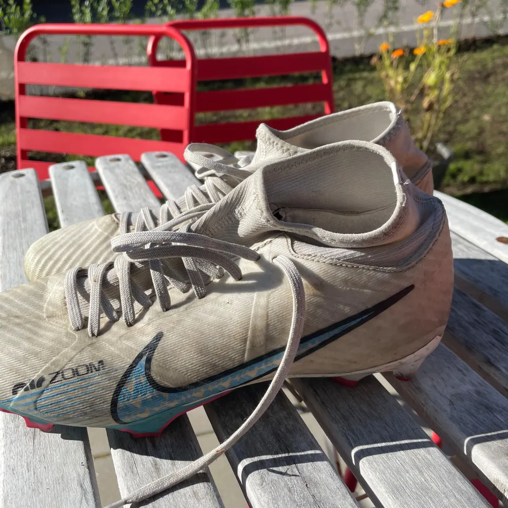 Säljer ett par Nike Mercurial fotbollsskor i en stilren beige färg med blå och röda detaljer. Skorna har snörning och är designade för att ge bra grepp på planen. De är i välanvänt skick men fortfarande funktionella för träning eller match.. Skor.